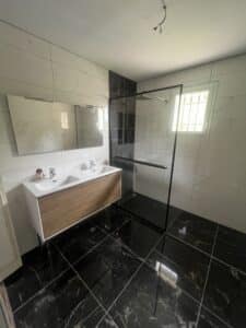 Salle de bain moderne : double vasque en bois, douche italienne noire, murs carrelés blancs et sol marbre noir. Miroir éclairé, fenêtre à barreaux.
