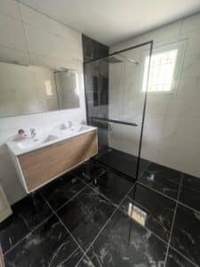 Salle de bain moderne avec double vasque bois, douche italienne à cadre noir, carrelage mural blanc marbré et sol noir veiné.
