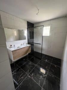 Salle de bain moderne. Sol marbre noir brillant, murs carrelage blanc. Double vasque bois, miroir. Douche italienne cadre noir. Fenêtre.