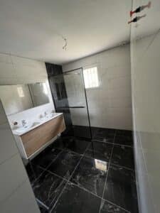 Salle de bain moderne rénovée. Carrelage noir marbré au sol, blanc aux murs. Double vasque bois, grande douche italienne à cadre noir et miroir.